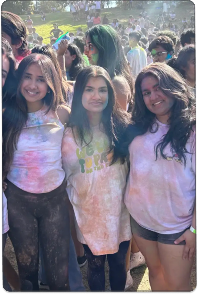 A group of young people pose together in a crowded outdoor setting, with colorful powder visible on their clothes and faces.