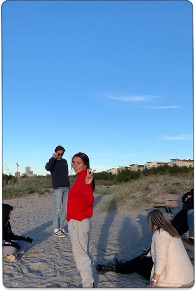 A group of young people spend time together on a sandy beach or dune area under a clear blue sky, with several people standing and sitting in the background.